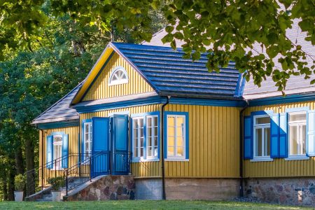 A traditional yellow wooden house with blue window shutters and a stone foundation, surrounded by trees and greenery, with sunlight casting shadows on the facade.