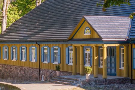 A traditional yellow wooden house with blue window shutters and a stone foundation, surrounded by trees and greenery, with sunlight casting shadows on the facade.