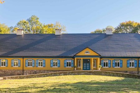 A traditional yellow wooden house with blue window shutters and a stone foundation, surrounded by trees and greenery, with sunlight casting shadows on the facade.