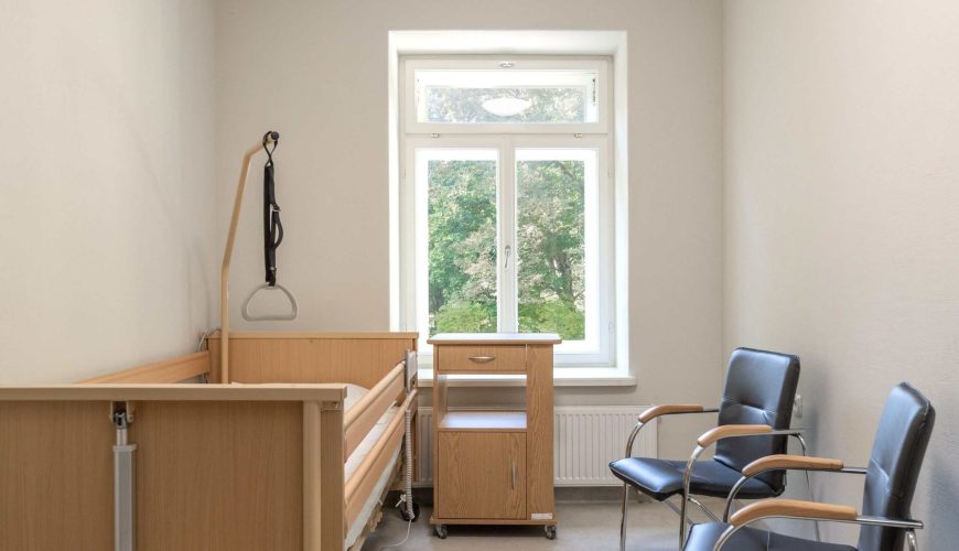 A small, bright hospital room with a wooden hospital bed, a bedside table, and two black chairs by a large window showing green trees outside. The walls are plain white, and the room appears clean and minimalistic.