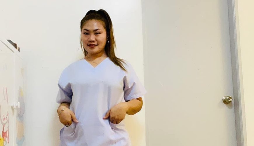 A woman wearing light purple scrubs and white sneakers stands indoors on a wooden floor, posing with her hands in her pockets in front of a white door and wall.