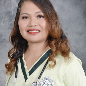 A woman with long wavy hair wearing a light green medical uniform, a stethoscope around her neck, and a badge on her chest, smiling in front of a gray background.