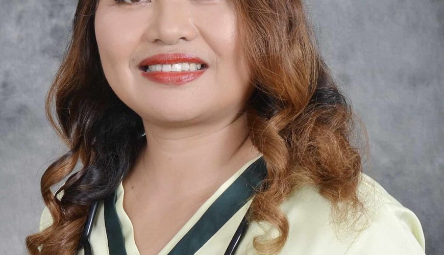 A woman with long wavy hair wearing a light green medical uniform, a stethoscope around her neck, and a badge on her chest, smiling in front of a gray background.