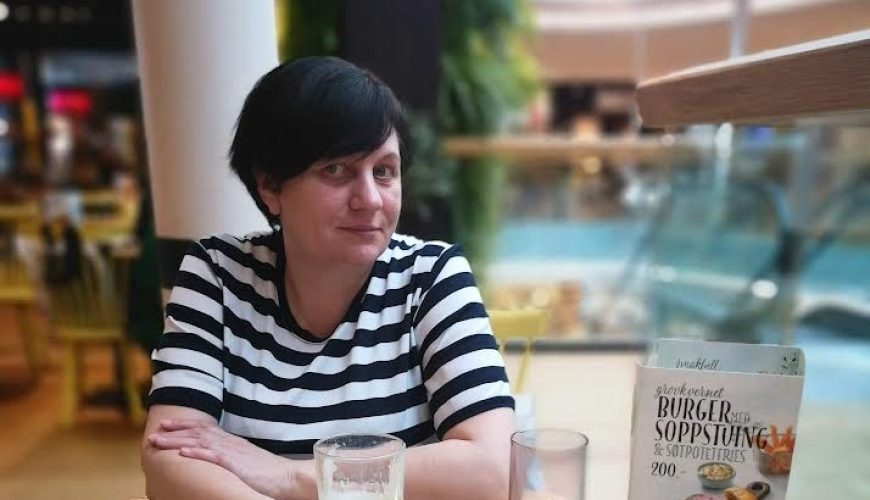 A person with short dark hair and a striped shirt sits at a wooden table in a restaurant with two glasses of latte, a candle, a menu, and a black wallet on the table. The background shows a bright, modern dining area.