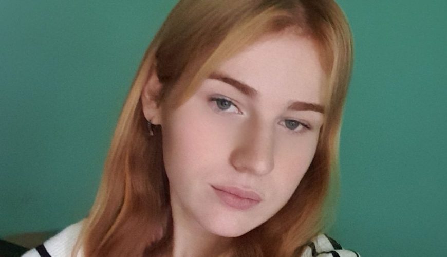 A young woman with long, light brown hair wearing a white and black striped sweater poses for a selfie in front of a teal wall. She has a neutral expression and is looking directly at the camera.