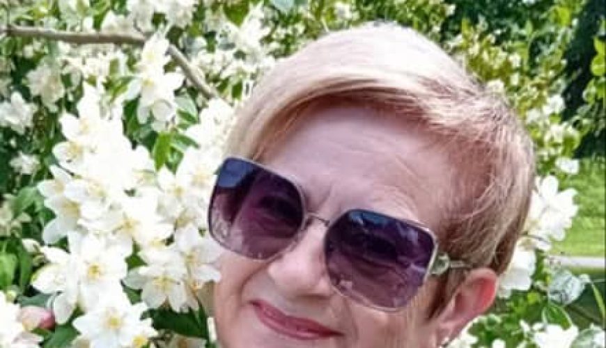 A smiling older woman with short blonde hair and sunglasses stands in front of blooming white flowers and green foliage on a sunny day. She is wearing a patterned top and a dark vest.