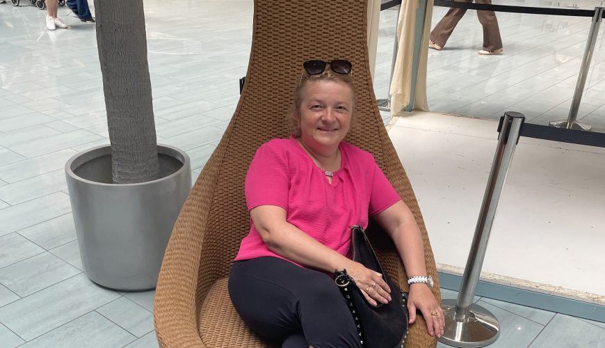 A woman in a pink top and dark pants sits on a wicker chair, smiling. She has sunglasses on her head and is holding a black bag. The setting appears to be indoors with tiled floors and a large potted plant nearby.
