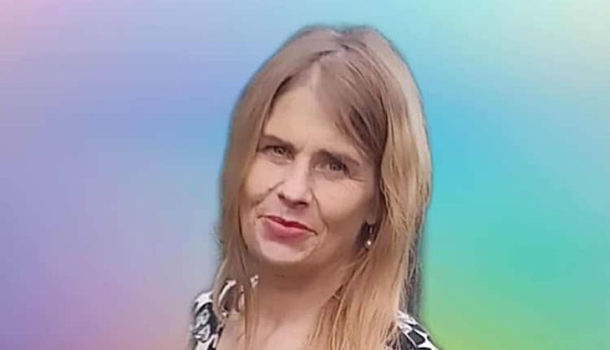 A woman with long blonde hair wearing a black and white floral top stands in front of a soft, pastel-colored gradient background, smiling gently at the camera.