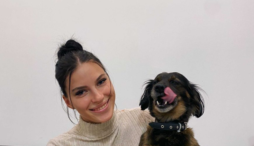 A woman with dark hair in a bun smiles while holding a small brown dog that is licking its nose, sitting on a gray couch against a plain white wall.