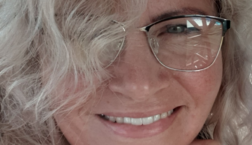 A person with wavy blonde hair, glasses, and a black shirt smiles at the camera, resting their chin on their hand. The background is softly blurred with some greenery and blue sky visible.
