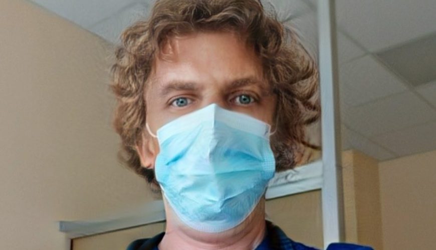 A person wearing blue scrubs, a surgical mask, and a stethoscope stands indoors, possibly in a hospital or clinic, looking directly at the camera.