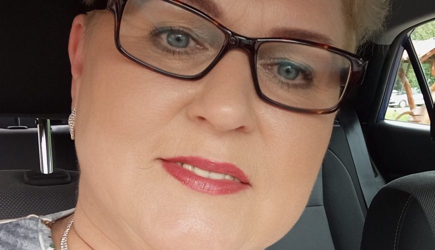 A woman with short blonde hair and glasses smiles slightly while sitting in a car. She is wearing a floral top, silver earrings, and a sparkly necklace.