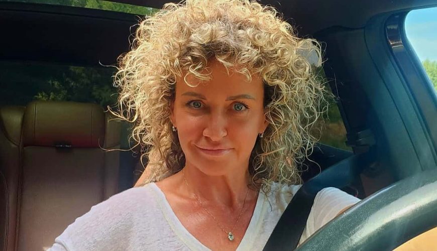 A woman with curly blonde hair and a white shirt sits in the driver's seat of a car, smiling at the camera with sunlight streaming through the open sunroof.