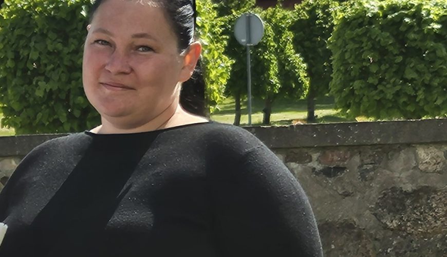 A woman with dark hair pulled back, wearing a black top, stands outdoors in front of a stone wall and green trees on a sunny day. She is smiling slightly and looking at the camera.