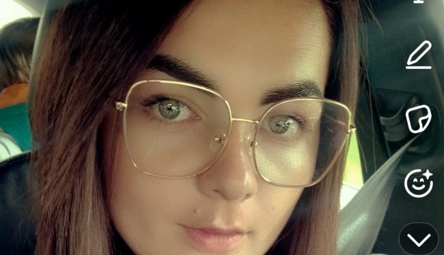 A woman with straight brown hair, wearing large transparent glasses and a gray hoodie, takes a selfie in a car. She has a seatbelt on, and the Snapchat app interface is visible on the screen.