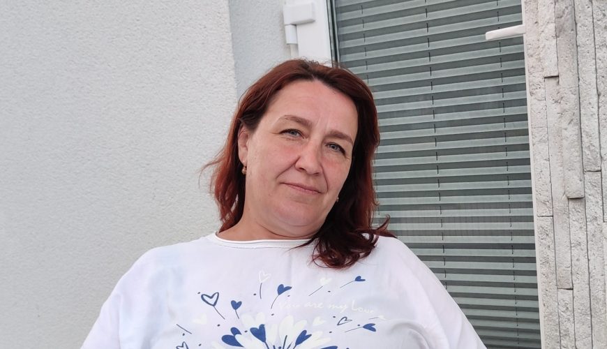A woman with reddish-brown hair sits on a chair outdoors, wearing a white shirt with a blue floral design and light blue pants. She is relaxed, with one hand resting on her lap, next to a white wall and closed blinds.
