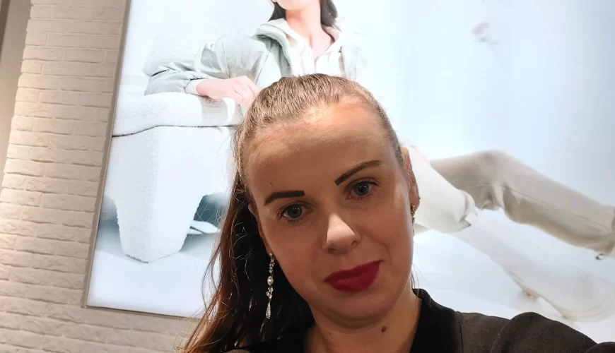 A woman with a ponytail and red lipstick takes a selfie indoors, standing in front of a large, bright poster featuring a model lounging in light-colored clothing and a cap.