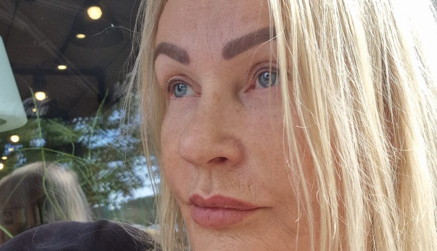 A woman with long blonde hair and arched eyebrows looks thoughtfully into the distance, sitting indoors near a window. She is wearing a dark top and natural light illuminates her face.