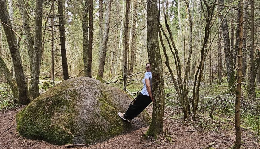 A person leans back against a thin tree trunk next to a large mossy rock in a dense forest, surrounded by tall trees and scattered branches on the forest floor.