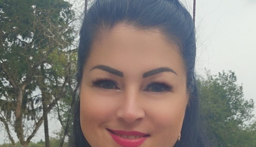 A woman with long black hair and wearing a white lace top smiles at the camera outdoors, with trees and greenery in the background. She has sunglasses resting on her head.