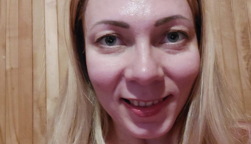 A woman with long blonde hair and light eyes smiles at the camera. She is wearing a pink top and is standing in front of a wooden plank wall.