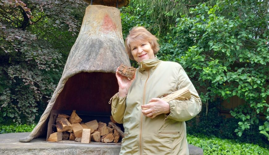 An older woman in a beige jacket stands by an outdoor stone fireplace, holding pieces of firewood and smiling. The fireplace is filled with wood, and lush green trees and bushes surround the area.
