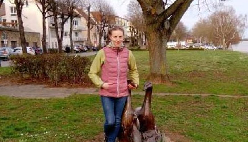 A person stands on grass next to a small animal sculpture under a large leafless tree, with residential buildings and a river visible in the background.