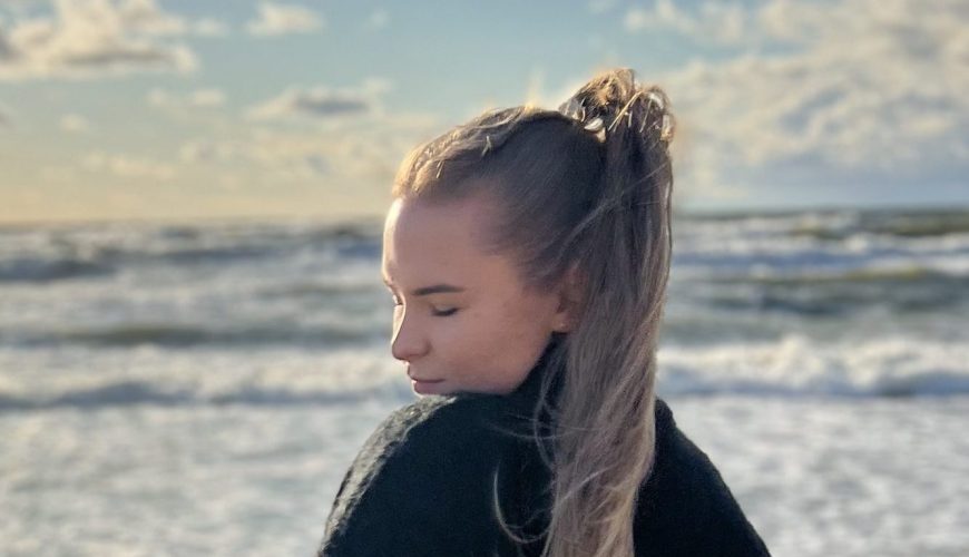 A woman in a black sweater and light jeans stands on a sandy beach, looking over her shoulder towards the ocean. The sun is low, casting a warm glow on the waves and the cloudy sky. She holds a small bag by her side.