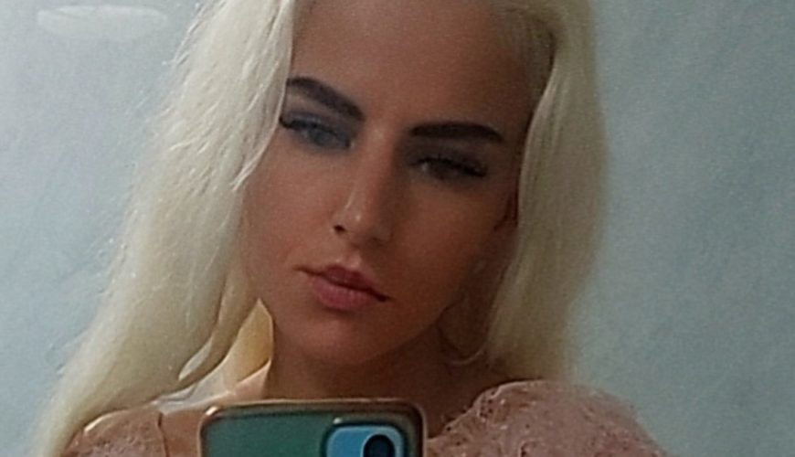 A woman with long, straight platinum blonde hair takes a mirror selfie with a blue phone. She is wearing a pale pink top and has a neutral facial expression. The background is a light gray tiled wall.