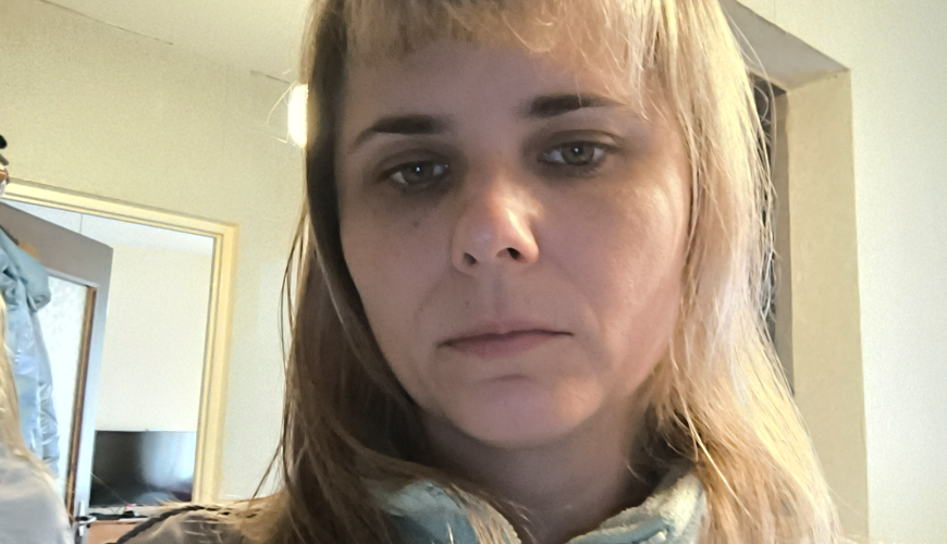 A woman with straight, light brown hair and bangs looks at the camera with a neutral expression. She is wearing a light blue zip-up top. The background shows a room with mirrors, a light, and a small red decoration.