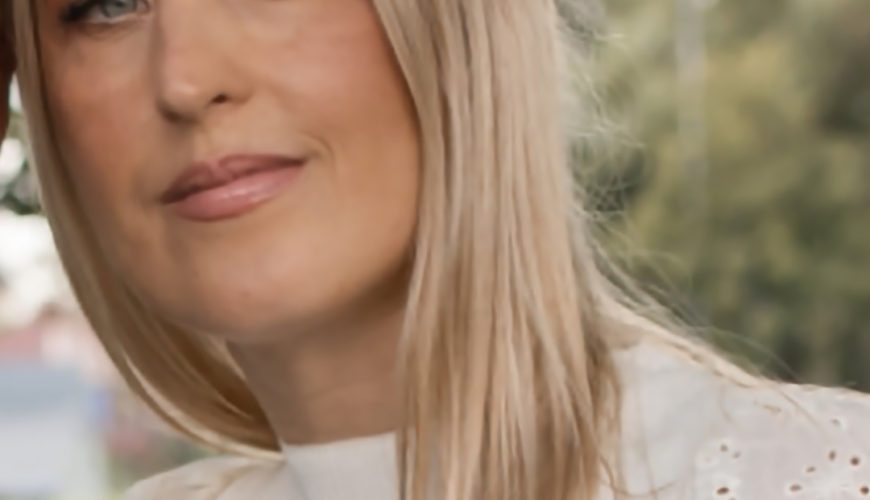 A woman with long, straight blonde hair and light skin wears a white eyelet top, standing outdoors with greenery blurred in the background.