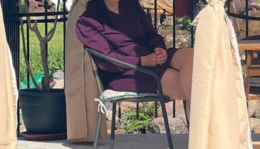 A woman with dark hair, wearing a purple outfit, sits and smiles on a chair under a gazebo with beige curtains. The background shows greenery, a fence, and outdoor structures in bright sunlight.