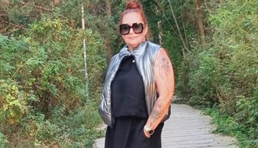 A person in a long black dress and silver vest stands on a wooden path surrounded by greenery, smiling and wearing sunglasses. It appears to be a sunny day in a natural outdoor setting.