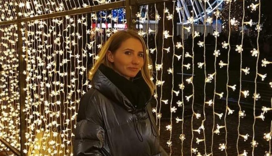 A woman in a long dark puffer coat stands under a tunnel of warm string lights at night, with a large illuminated Ferris wheel visible in the background.