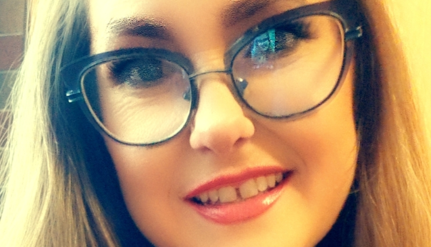A woman with long brown hair and glasses smiles at the camera. She is wearing a green top and a gold necklace. The background is softly blurred.