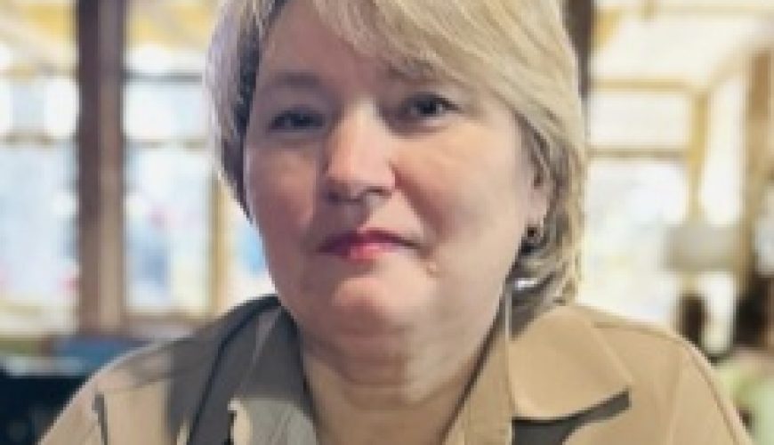 A woman with short, light blonde hair and a beige collared shirt sits indoors, facing the camera, with a blurred background of windows and warm lighting.