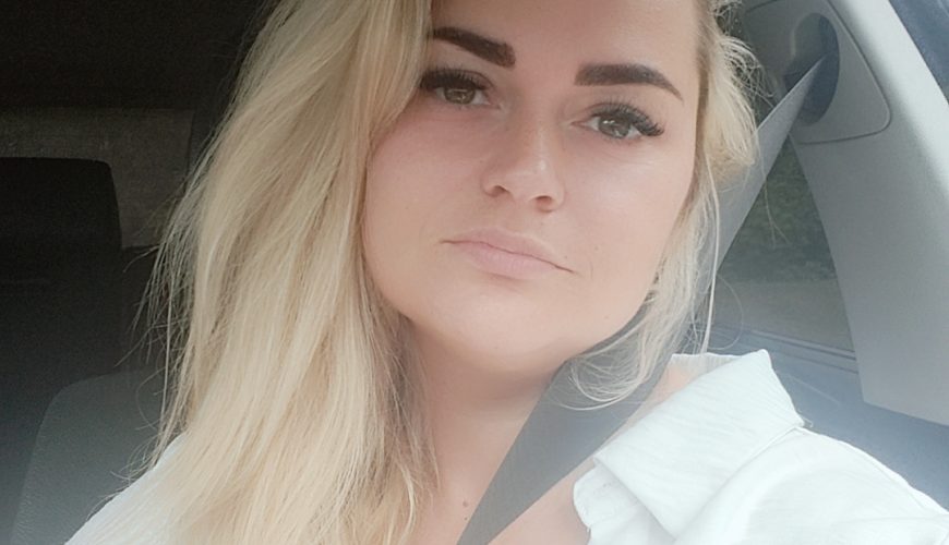 A woman with long blonde hair sits in a car wearing a white top and seatbelt, looking at the camera with a neutral expression. Natural light comes through the window.