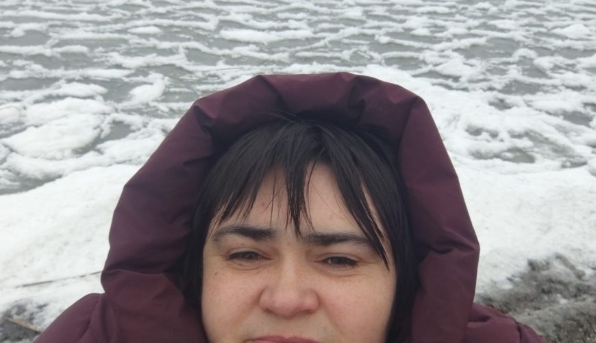 A person in a maroon hooded coat stands by a partially frozen lake, with patches of ice and snow on the water and cloudy skies overhead.