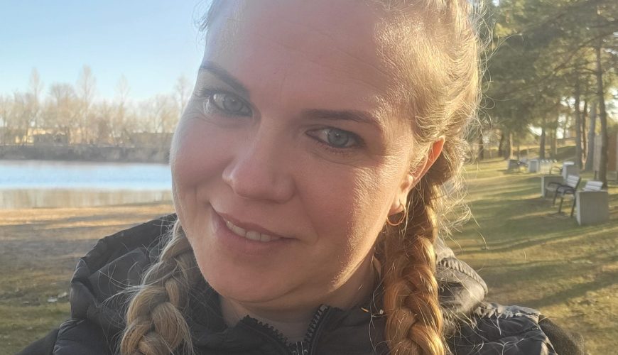 A woman with long blonde braids and a black jacket smiles at the camera outdoors on a sunny day, with trees, a lake, and park benches in the background.