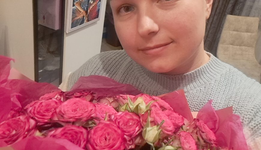 A person with blonde hair, wearing a gray sweater, holds a bouquet of vibrant pink roses wrapped in pink paper, smiling softly at the camera indoors.