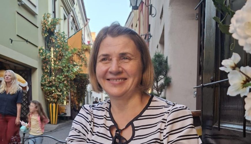 A woman in a striped shirt sits at a glass table outside a café on a narrow street, smiling. Flowers decorate the table; people walk in the background. The scene is bright and cheerful.