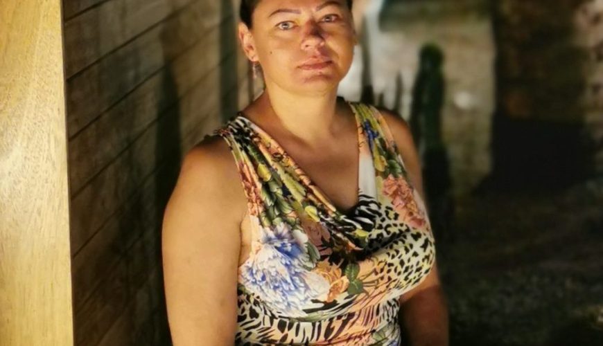 A woman in a patterned dress sits on a low wall at night, beside palm trees and cacti, with a wooden fence and a street in the background illuminated by streetlights.