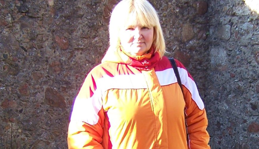 A woman with blonde hair, wearing a bright orange and red jacket and dark pants, stands with her hands in her pockets in front of a stone wall with a barred window in an outdoor, overgrown area.