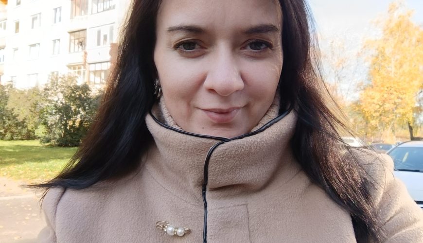 A woman with long dark hair wearing a beige coat with a pearl brooch stands outside on a sunny day, with a building, trees, and a parked car in the background.