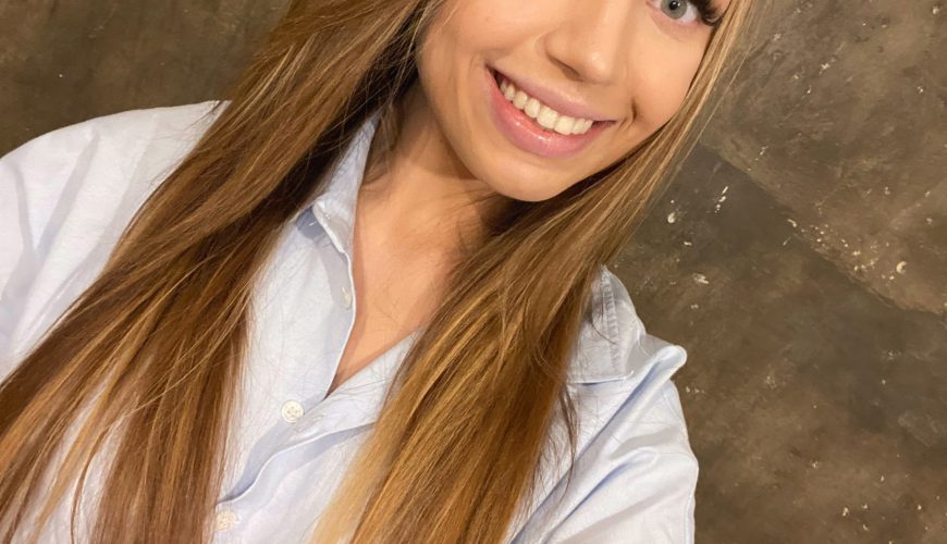 A young woman with long, straight blonde hair and a light blue shirt smiles at the camera, taking a selfie against a brown, textured wall with a visible electrical outlet.