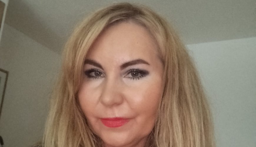 A woman with long blonde hair and red lipstick smiles softly while looking at the camera, standing indoors with a neutral background.