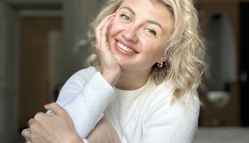 A smiling woman with wavy blonde hair, wearing a white sweater, sits indoors with her hand resting on her cheek. She looks at the camera in a well-lit room with neutral-colored walls and furniture in the background.