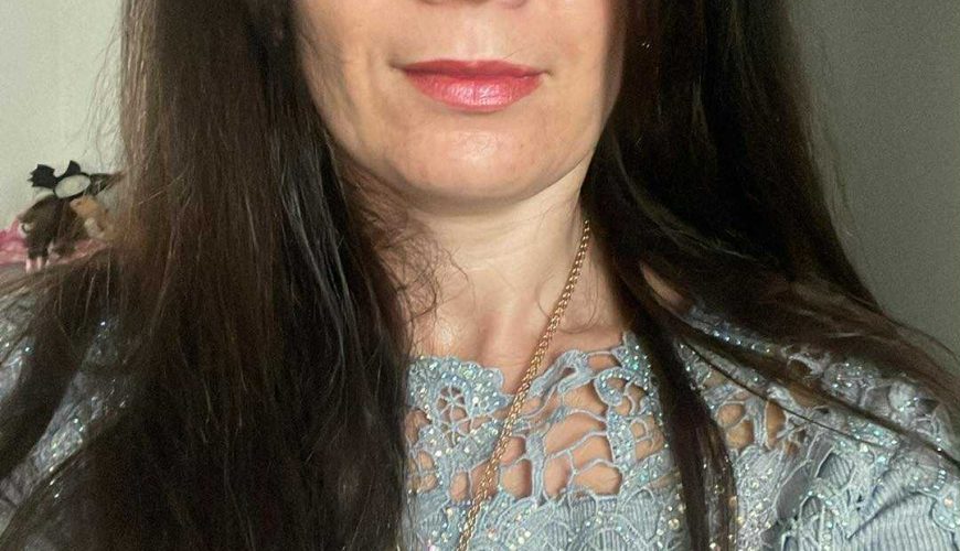 A woman with long brown hair wearing a light blue top with lace detailing, looking at the camera indoors. She has light makeup, a necklace, and a neutral facial expression.