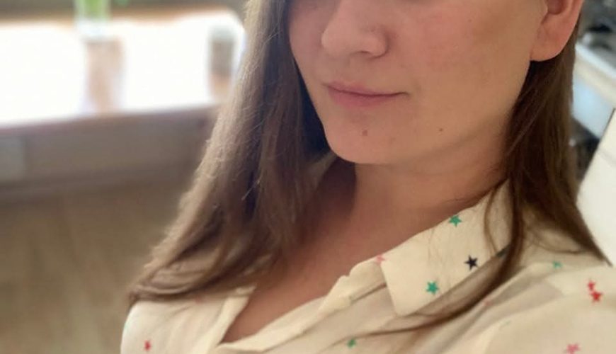 A woman with long brown hair and natural makeup looks at the camera, wearing a cream shirt with multicolored stars. She stands indoors, with a blurred bouquet of flowers on a table in the background.