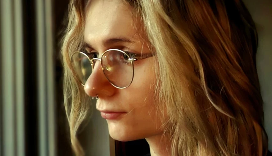 A person with long, wavy blonde hair and round glasses looks thoughtfully out of a window, with soft natural light illuminating their face.